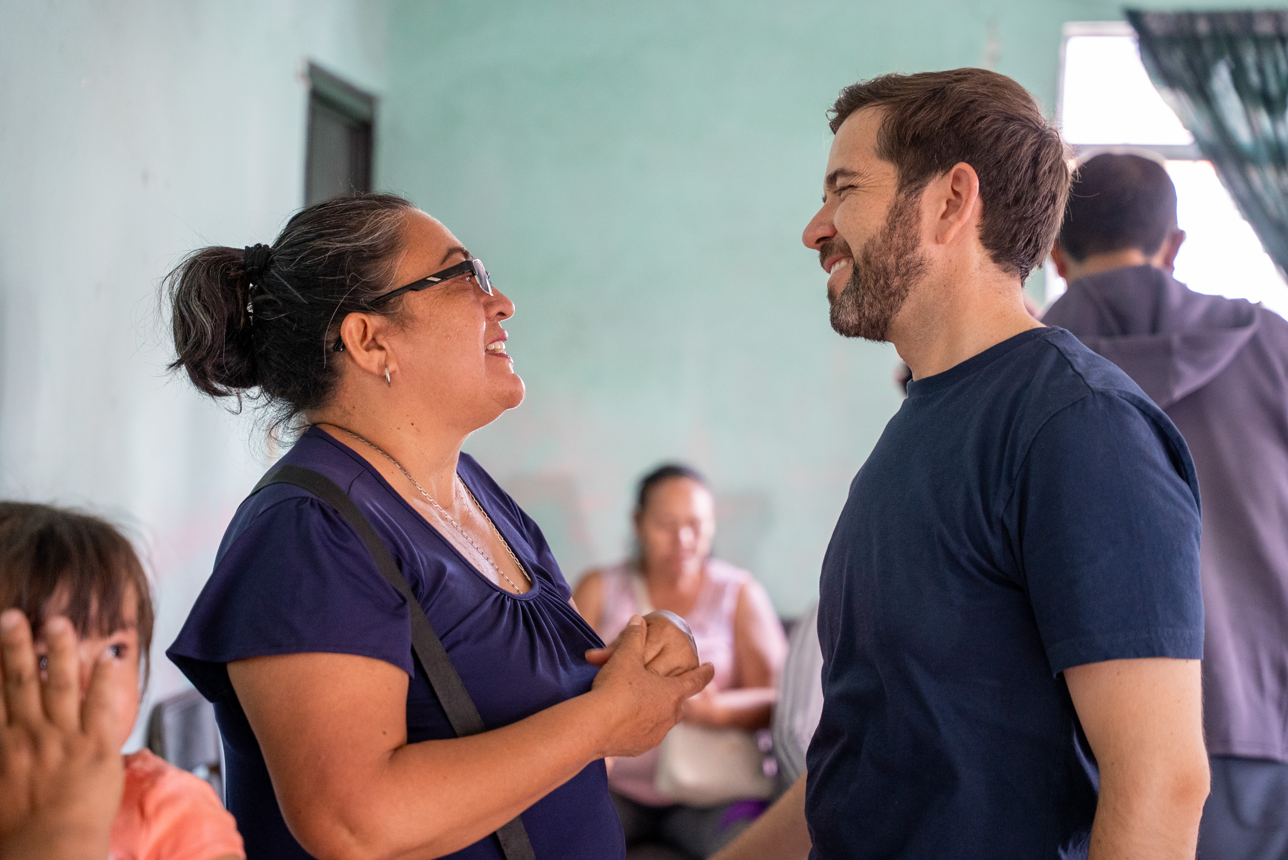 A smiling woman and man face each other and talk indoors with other people blurred in the background.