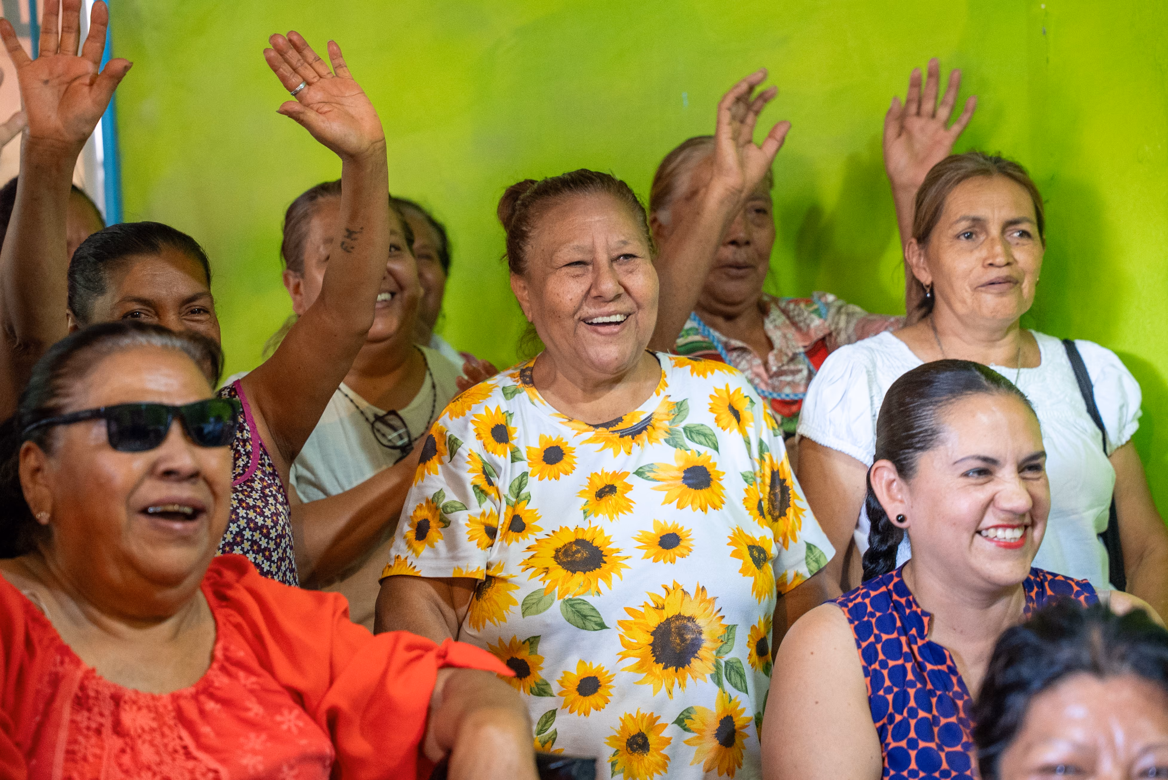 Group of smiling middle-aged women, some raising their hands, against a bright green wall.