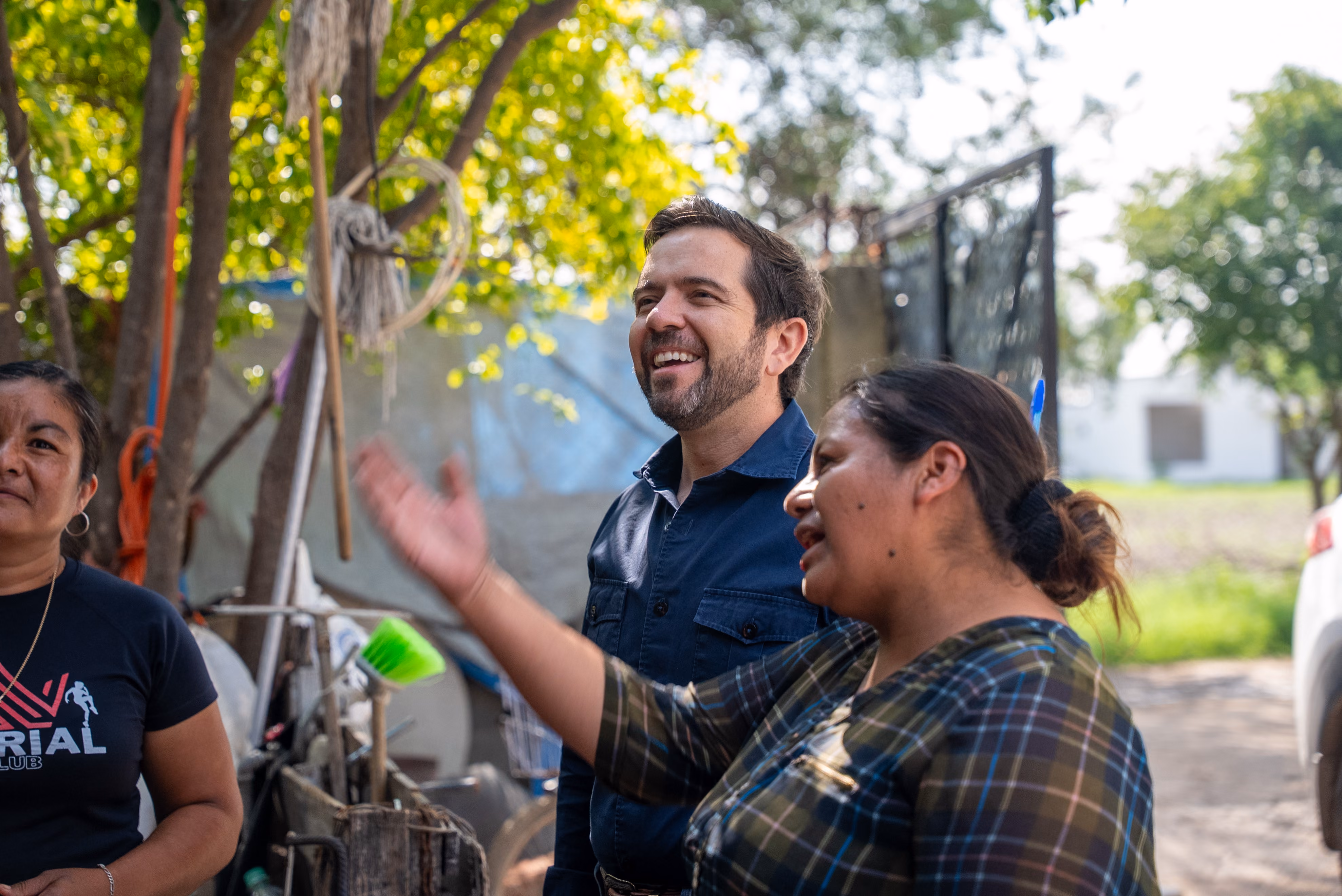 Three people outdoors engaged in friendly conversation near trees and a fence.