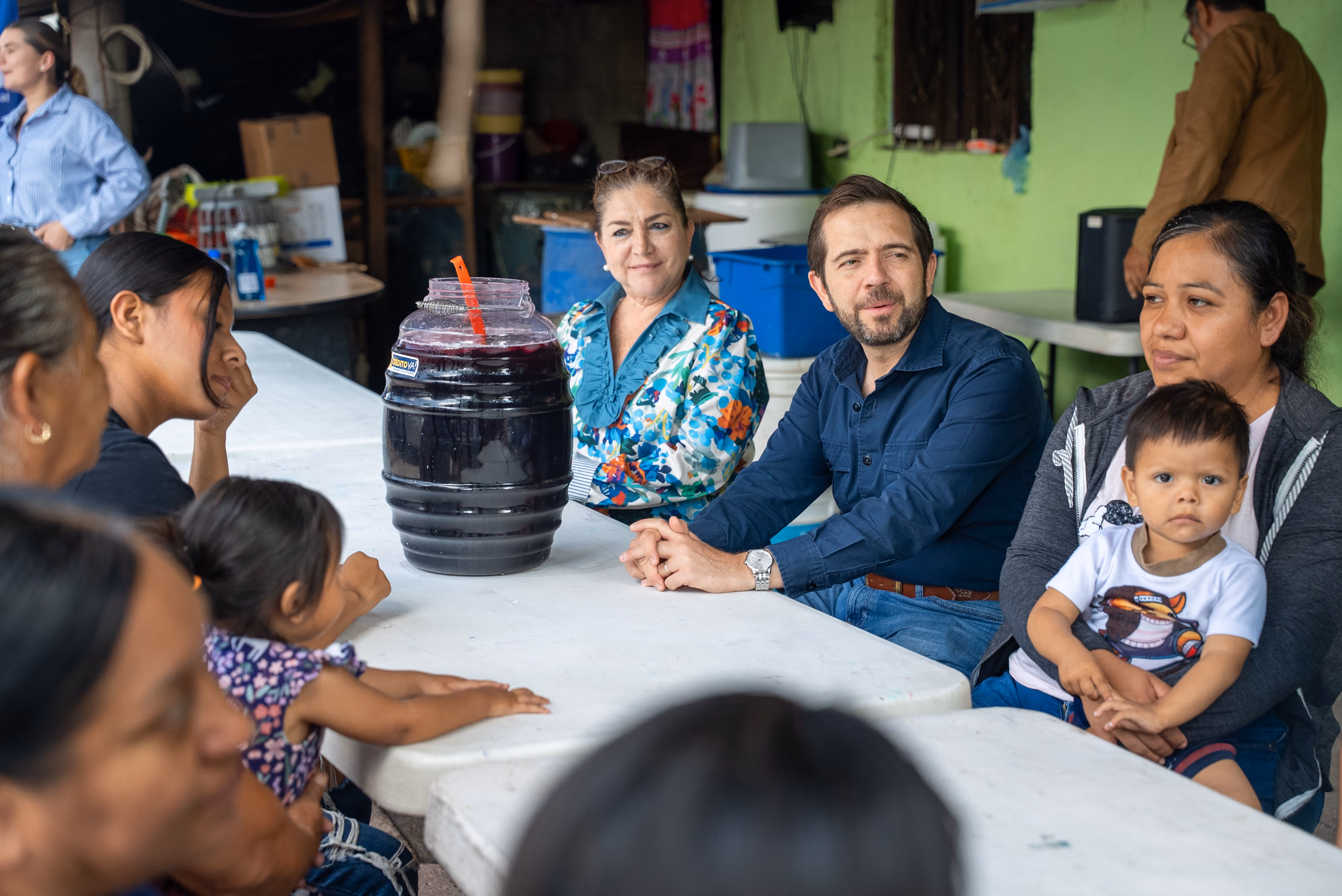 Group sitting around a table with a large jar of dark purple drink and a toddler on a woman's lap.
