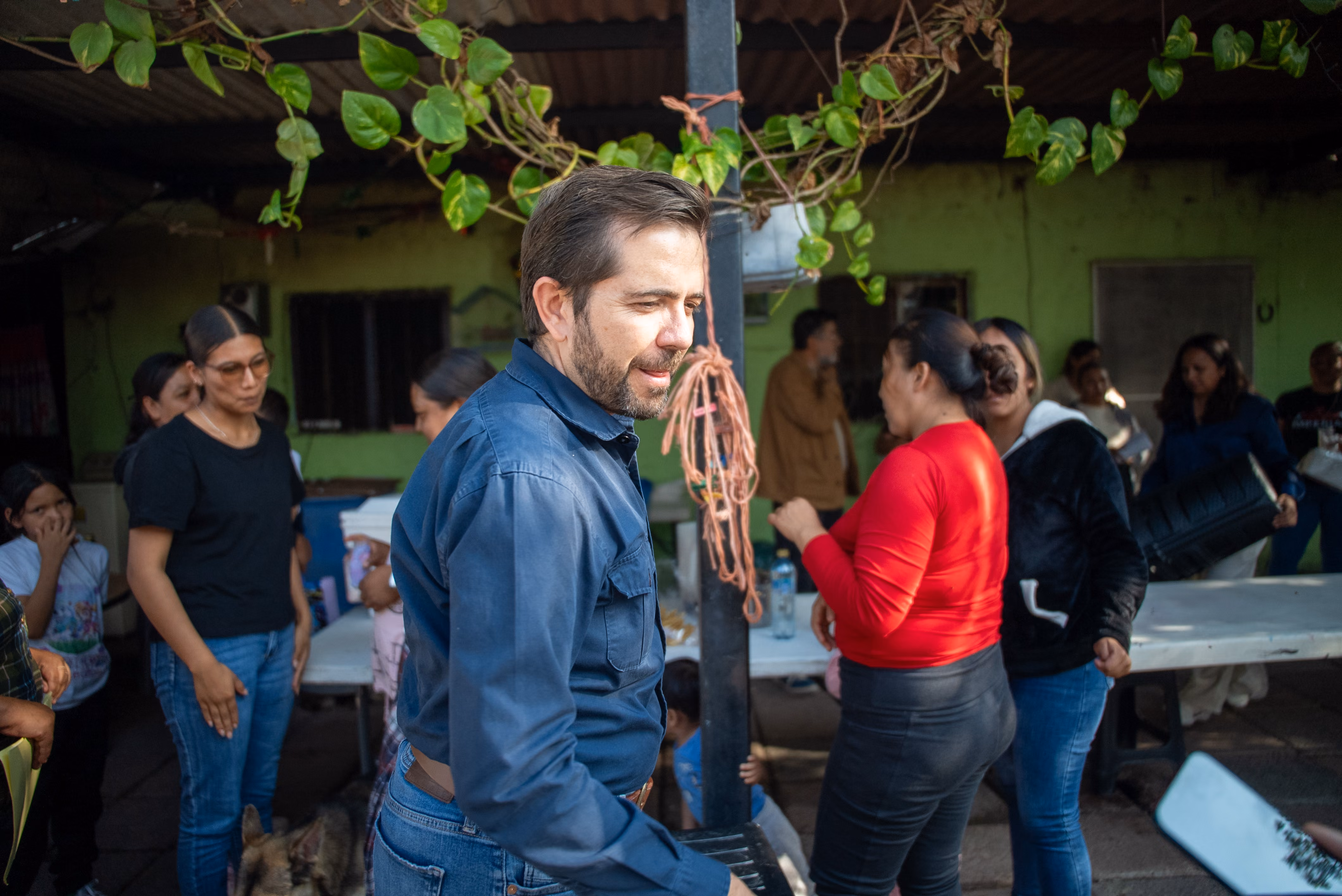Man in blue shirt among a group of people socializing in a shaded outdoor area with green walls.