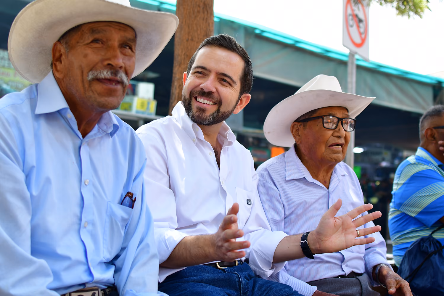 Three men sitting outdoors, two older men wearing white hats and a younger man smiling in the middle.