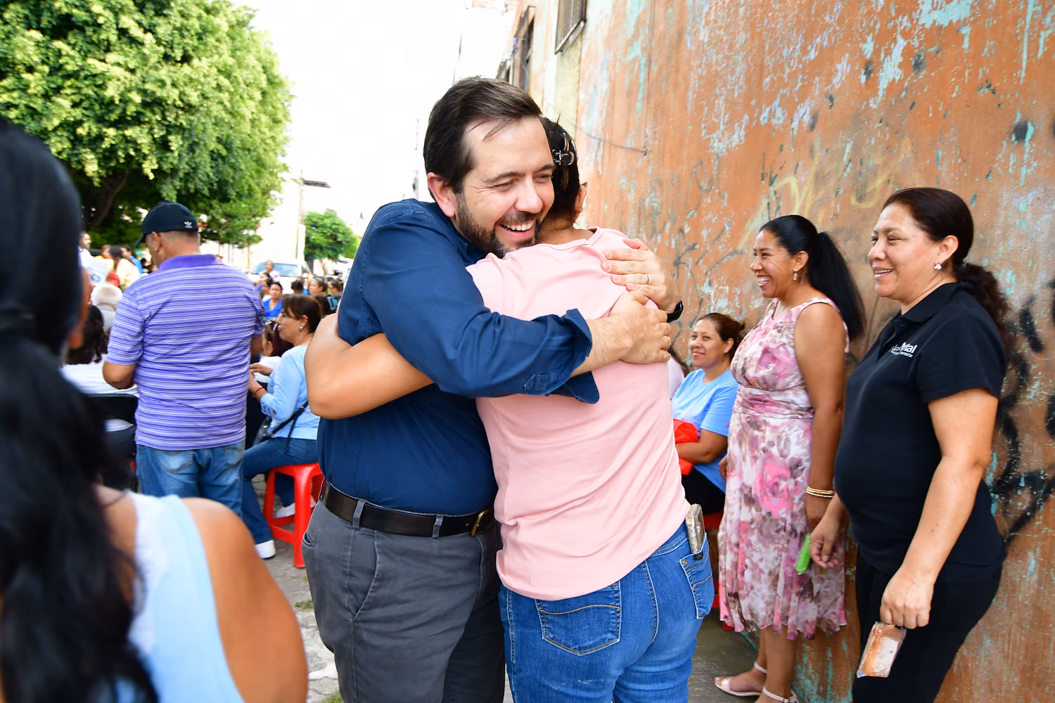 Man in blue shirt smiling warmly as he hugs a woman in a pink shirt outdoors with onlookers nearby.