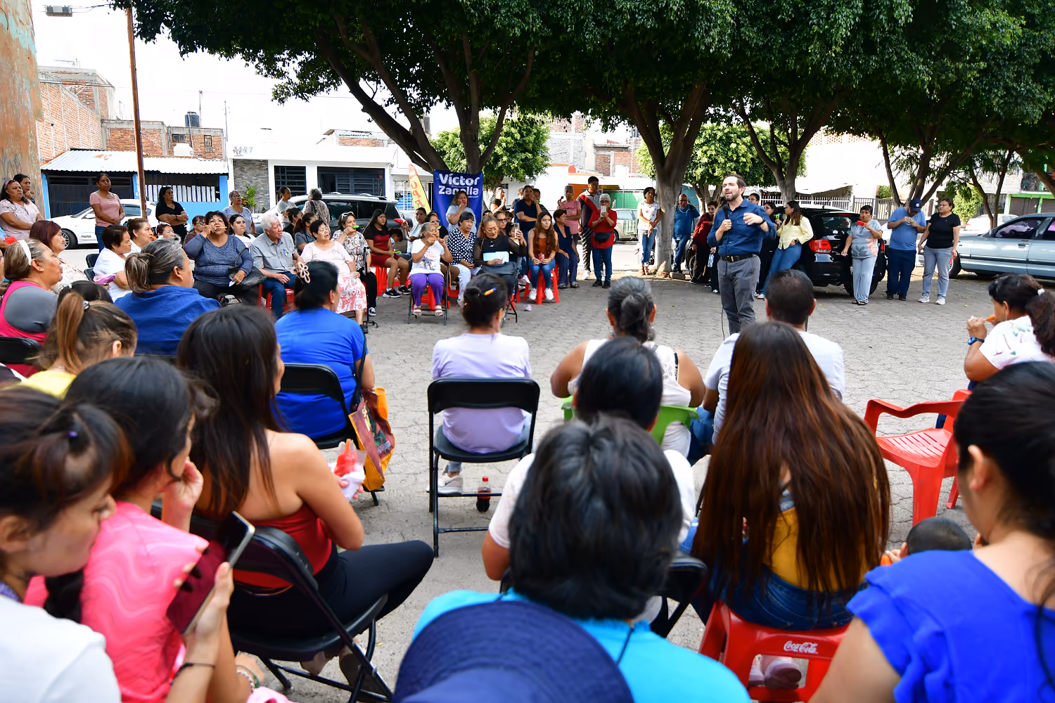 Man speaking with microphone to seated and standing crowd under trees in a neighborhood outdoor area.