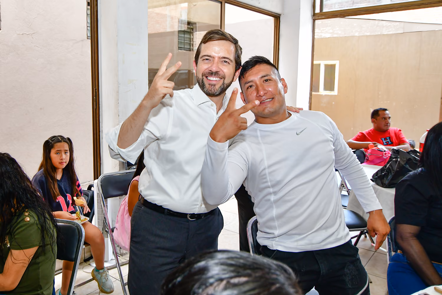 Two men smiling and showing peace signs with fingers in a room with seated people.