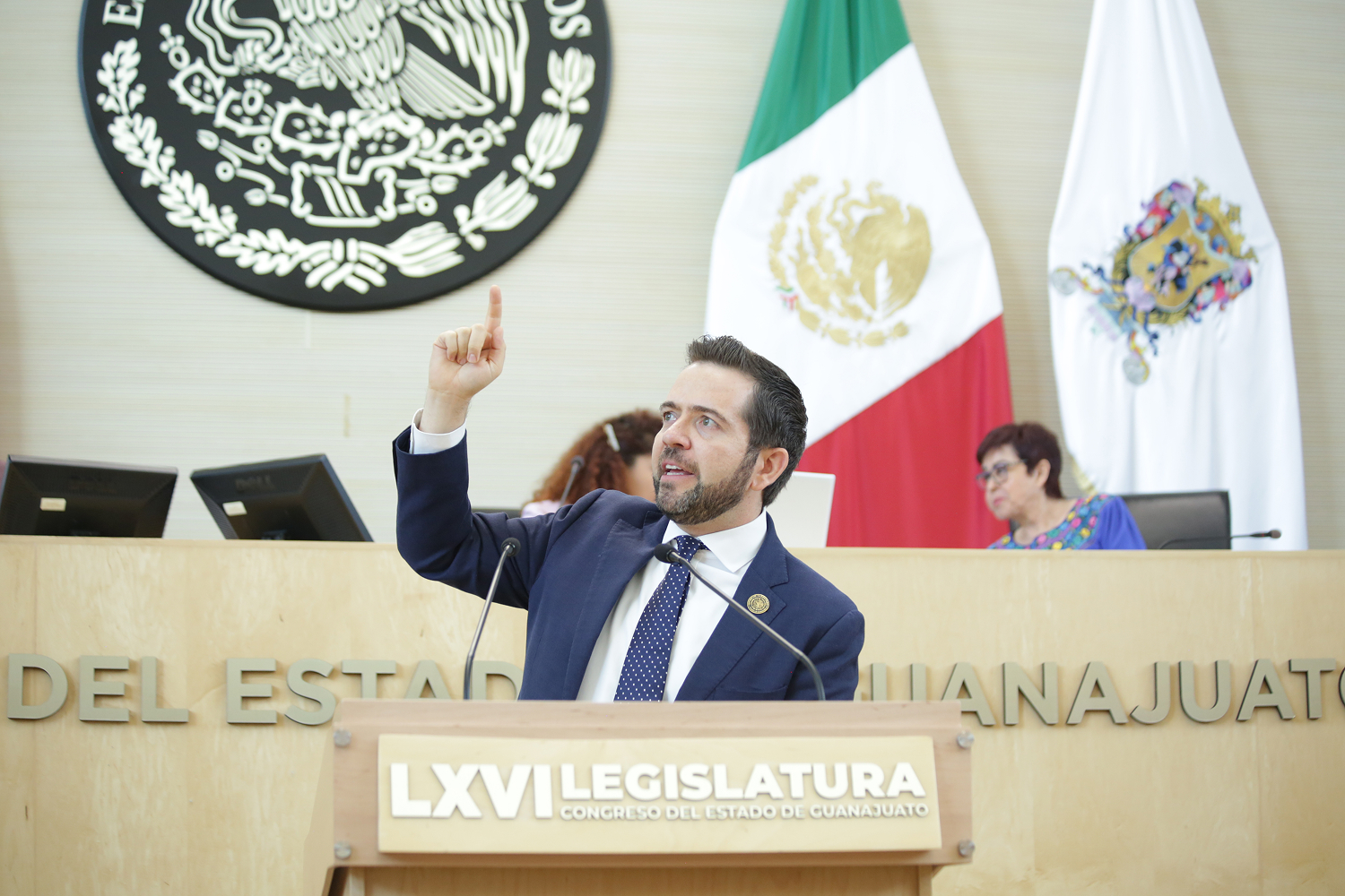 Empty legislative chamber with rows of seats and large Mexican flag behind the podium.