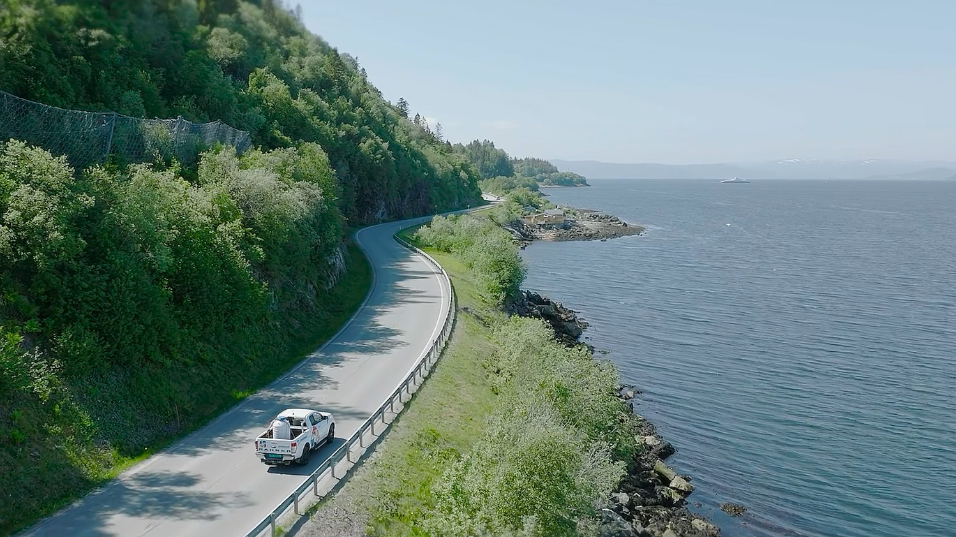 White pickup truck driving on coastal road beside green hills and a calm blue sea.