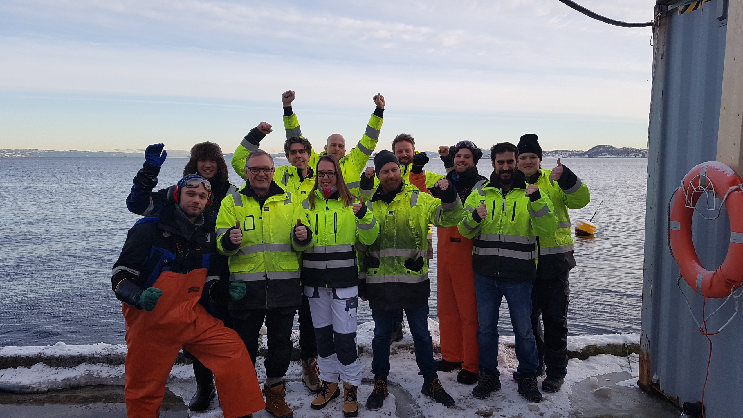 Group of people in high-visibility jackets cheering together by a snowy waterfront.