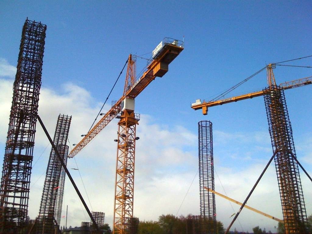 Tower cranes above a building site, with steel reinforcement cages for columns standing against a blue sky