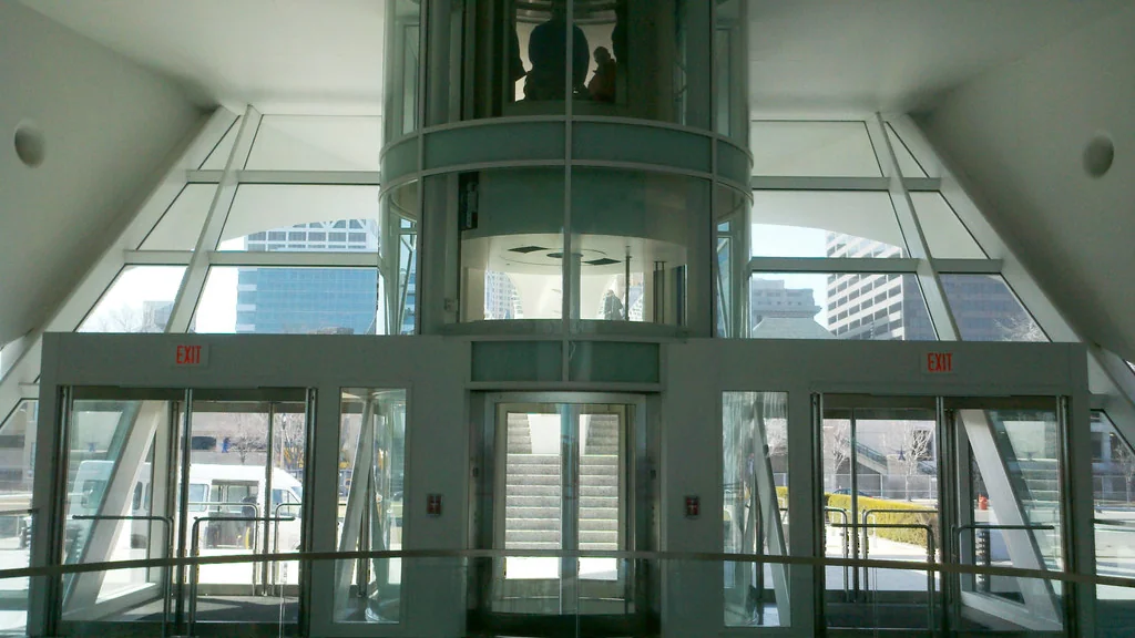 A round glass lift rising through a glazed building lobby, with a staircase and exit doors behind it