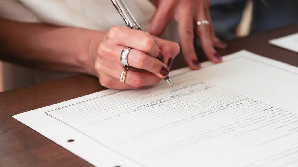 A hand signing a printed agreement with a pen at a desk