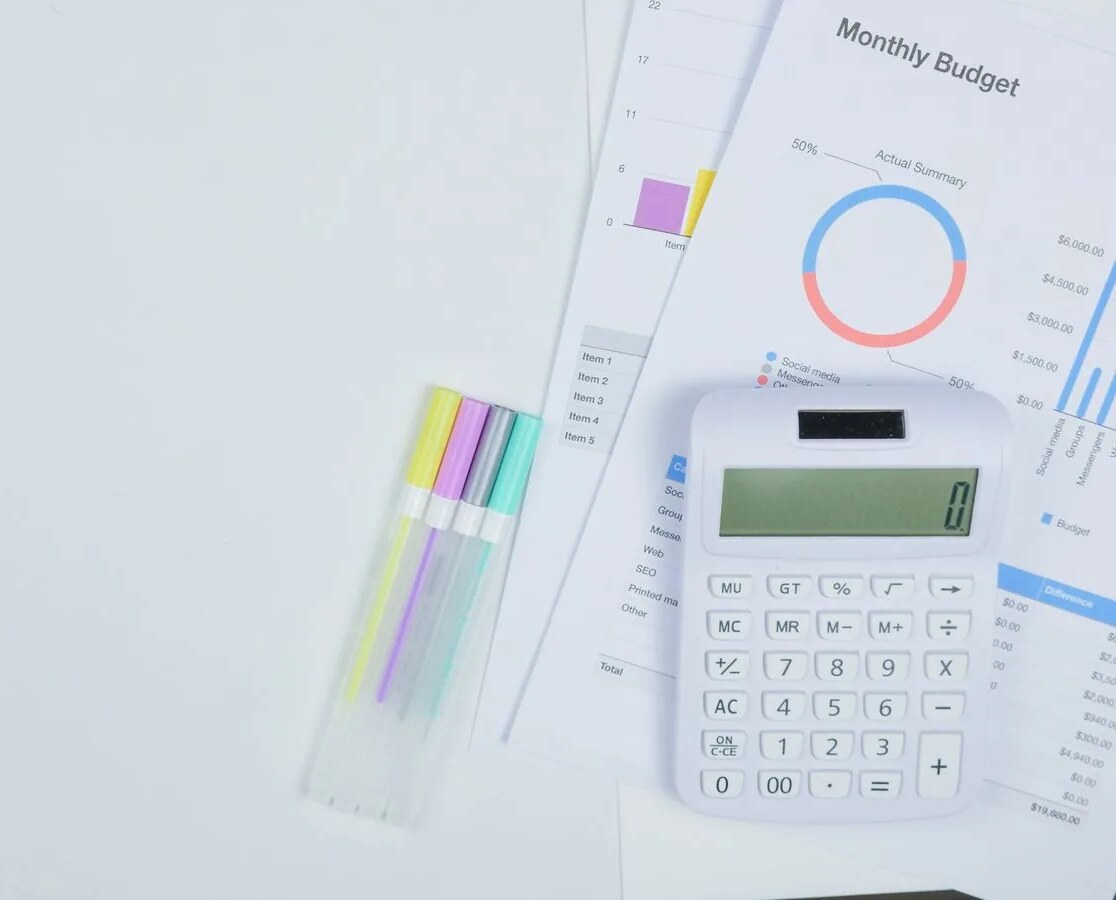 A monthly budget chart sheet with a calculator and a row of highlighters on a white desk
