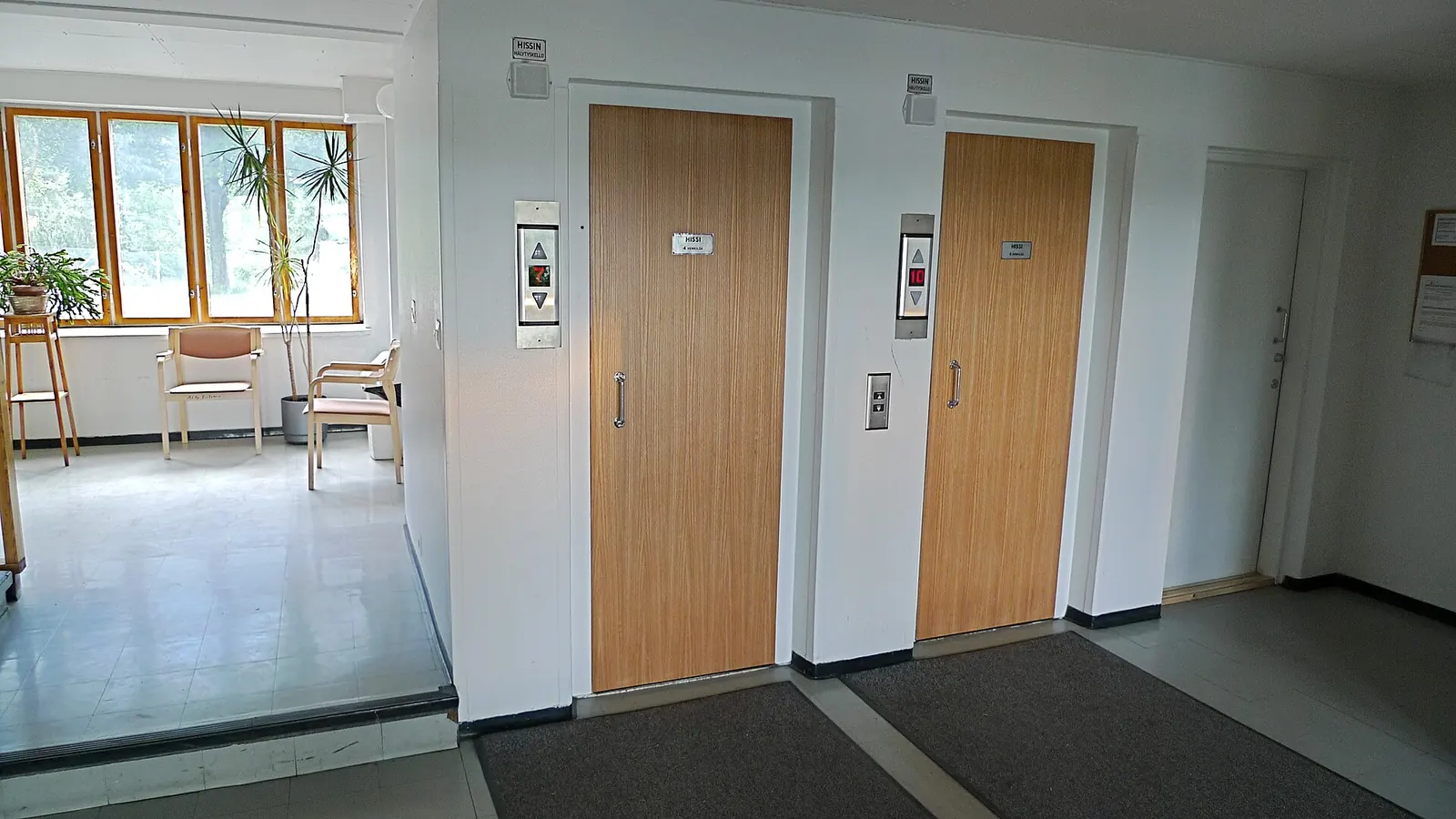 Two closed lift landing doors in a building lobby, each with a call panel and an alarm bell sign mounted above.