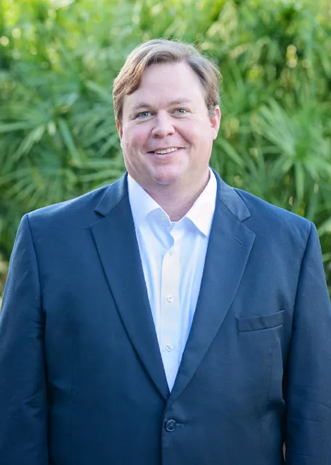 Smiling man in a dark suit jacket and white shirt standing outdoors with green plants behind him.