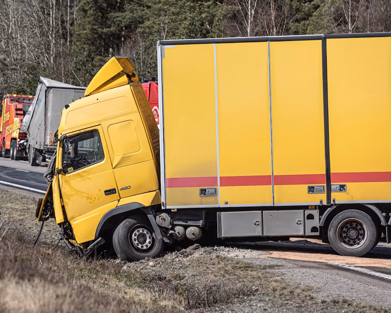 Yellow delivery truck with front damage stuck off the road beside a forested area.