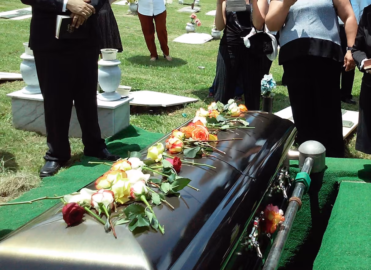 Black coffin with roses on top at a graveside service with people standing around in a cemetery.