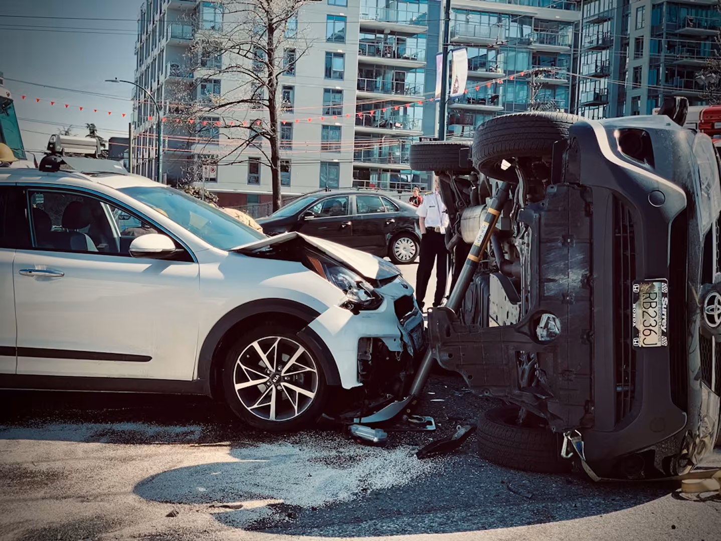 White car collided head-on with overturned black SUV on city street with building in background.