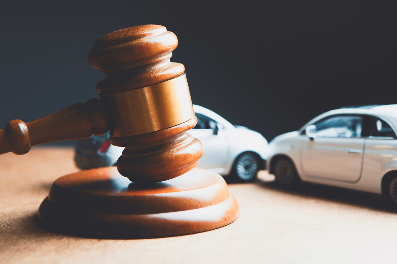 Wooden judge gavel with two white toy cars colliding in the background on a beige surface.