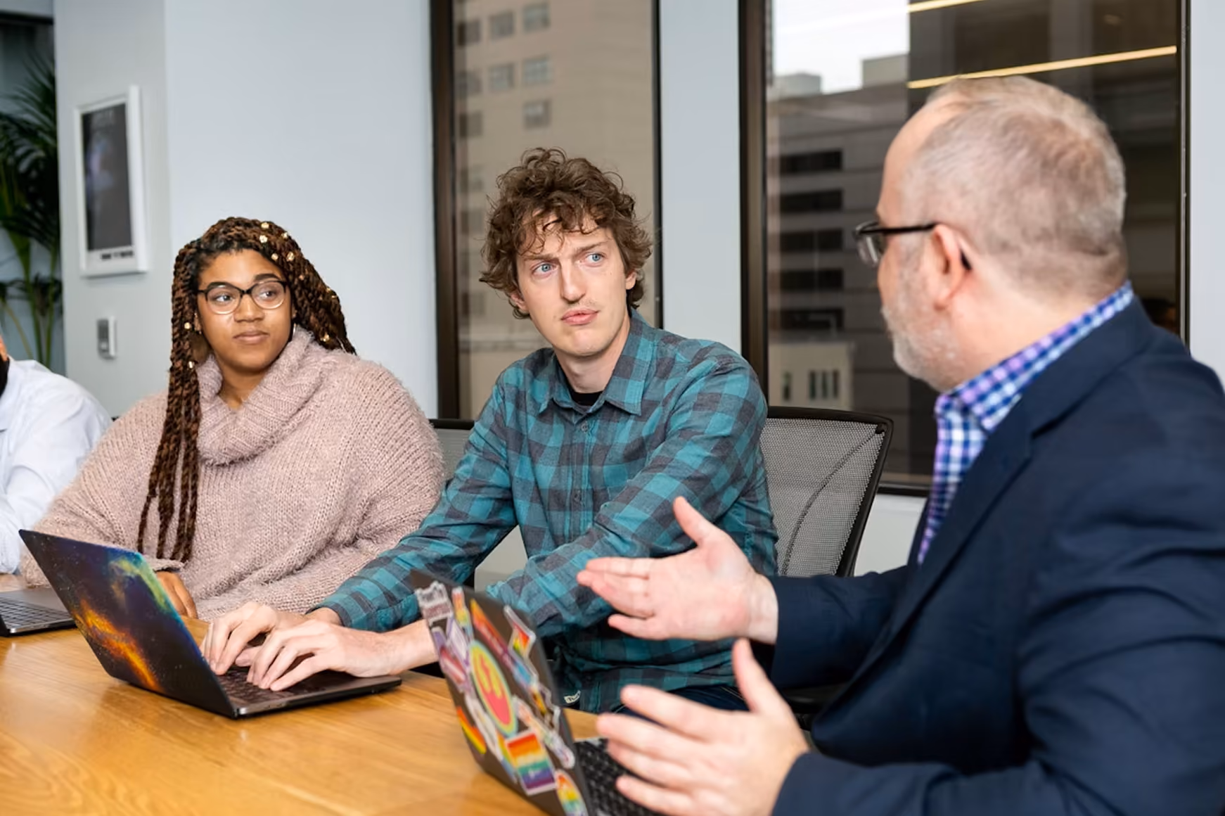 Three people in a meeting room with laptops, one man actively speaking and others listening.