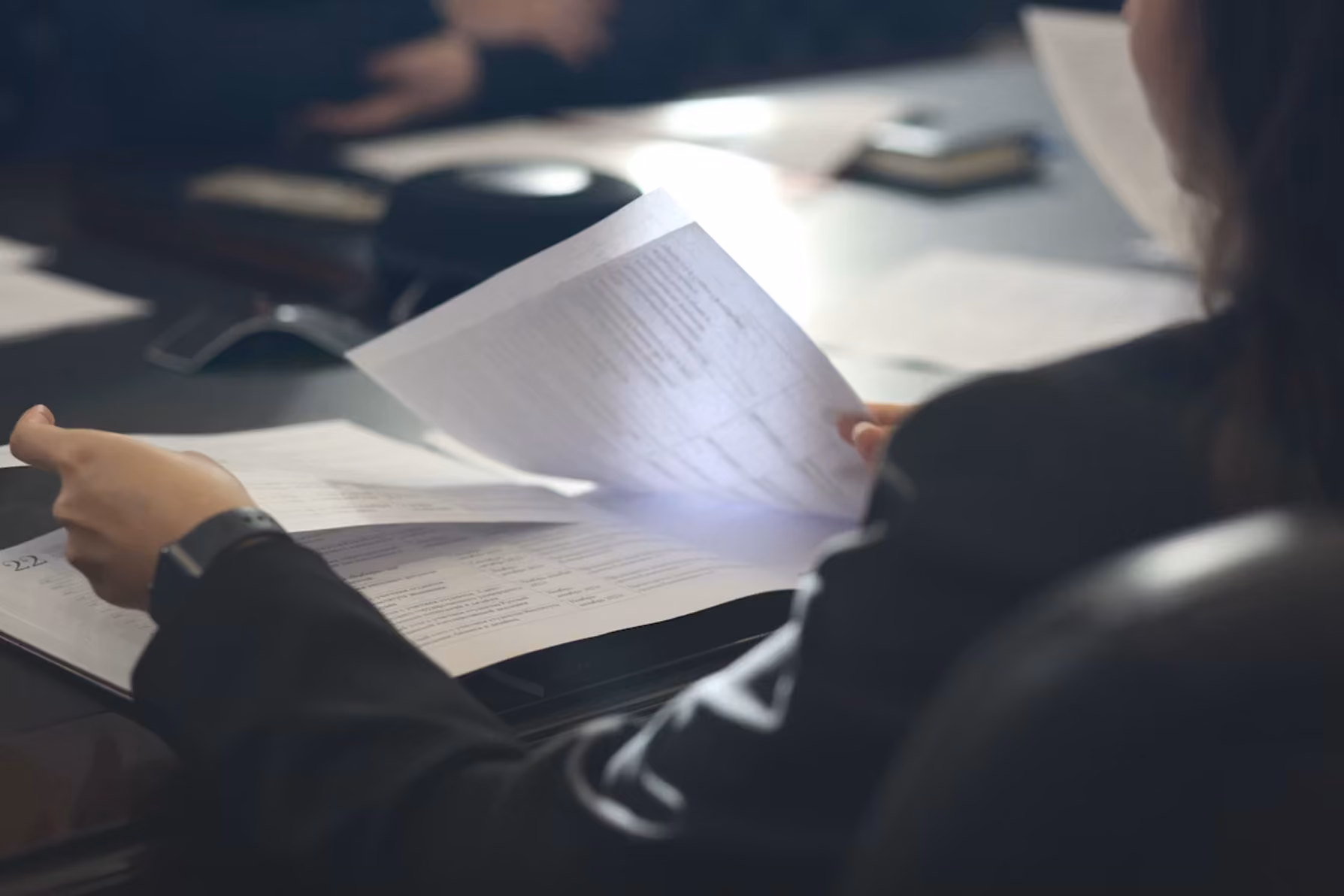 Person in dark clothing reading and turning pages of documents at a desk with blurred background.
