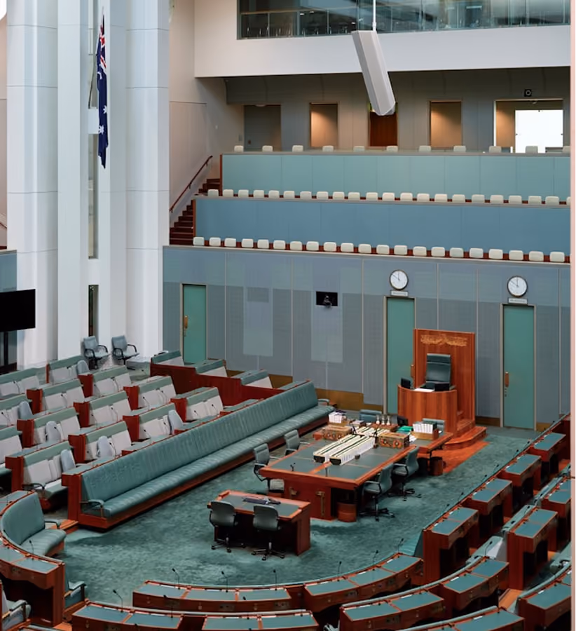 Empty parliamentary chamber with green seating, desks, and an Australian flag inside a modern building.