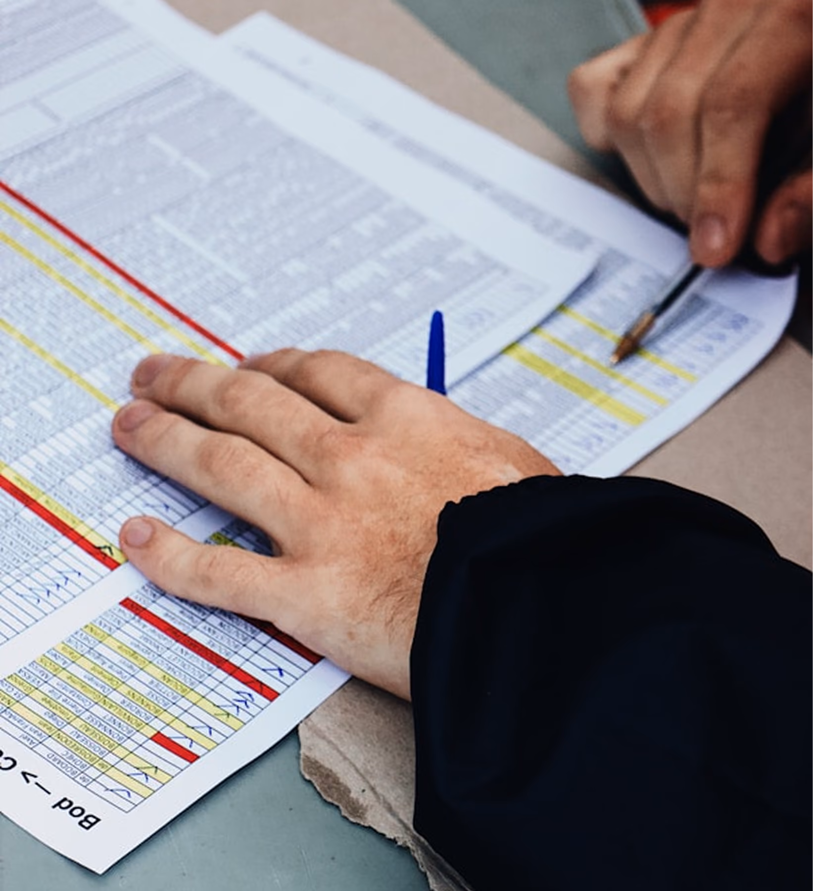 Two people reviewing printed tables or spreadsheets, one with a pen pointing at data rows in yellow and red.