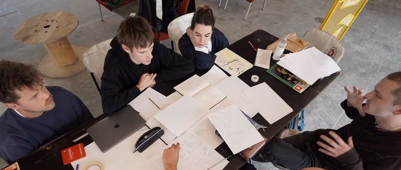 Four people sit around a table covered with papers, sketches, a laptop, and stationery in a work session.