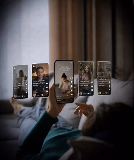 Person lying on couch holding phone with projected social media video thumbnails floating above.