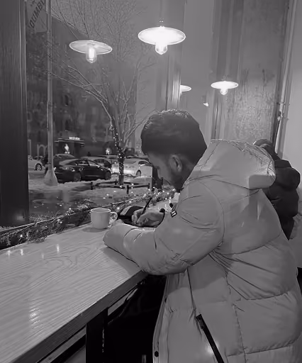Man in warm jacket writing at a cafe counter with city street visible through window at night.