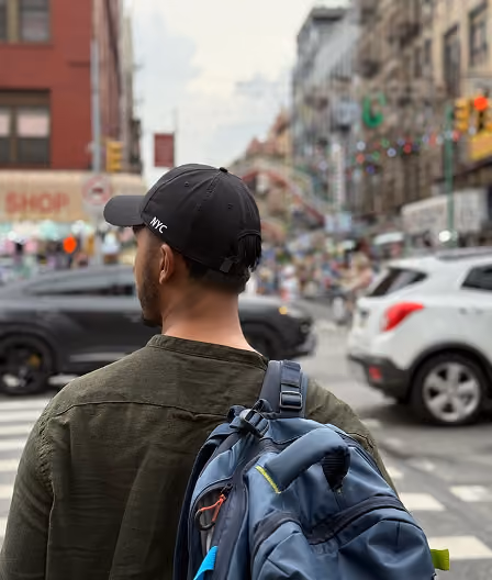 Man wearing black NYC cap and green jacket with a blue backpack standing on a city street.