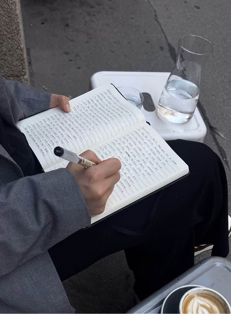 Person writing in a lined notebook with a pen while sitting outdoors near a glass of water and coffee.