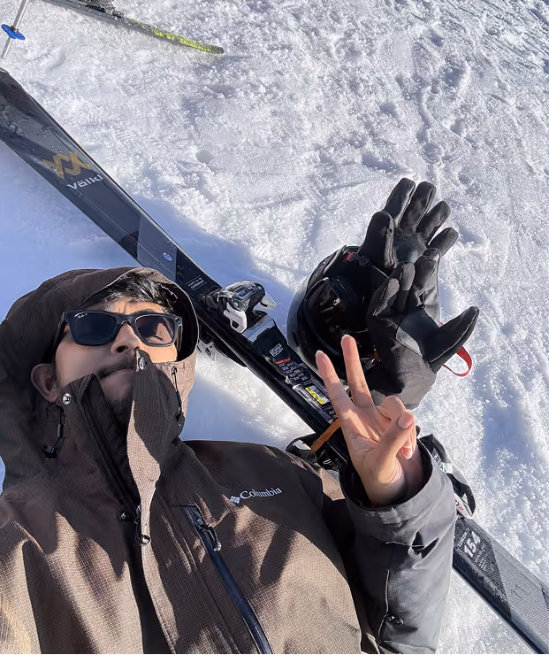 Person in sunglasses and brown jacket lying on snow showing a peace sign near skis and gloves.