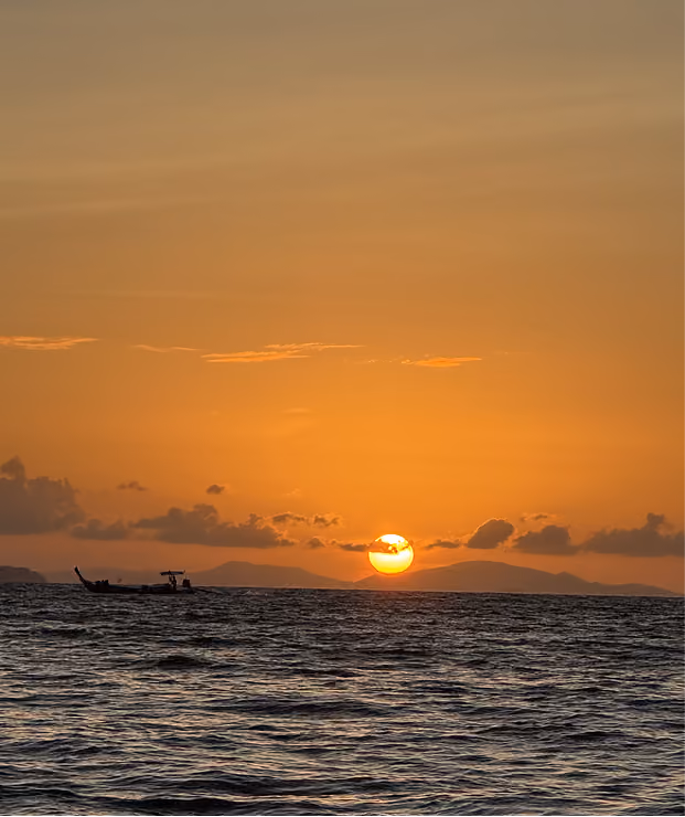 Sunset over the ocean with a small boat silhouetted near the horizon and distant mountains.