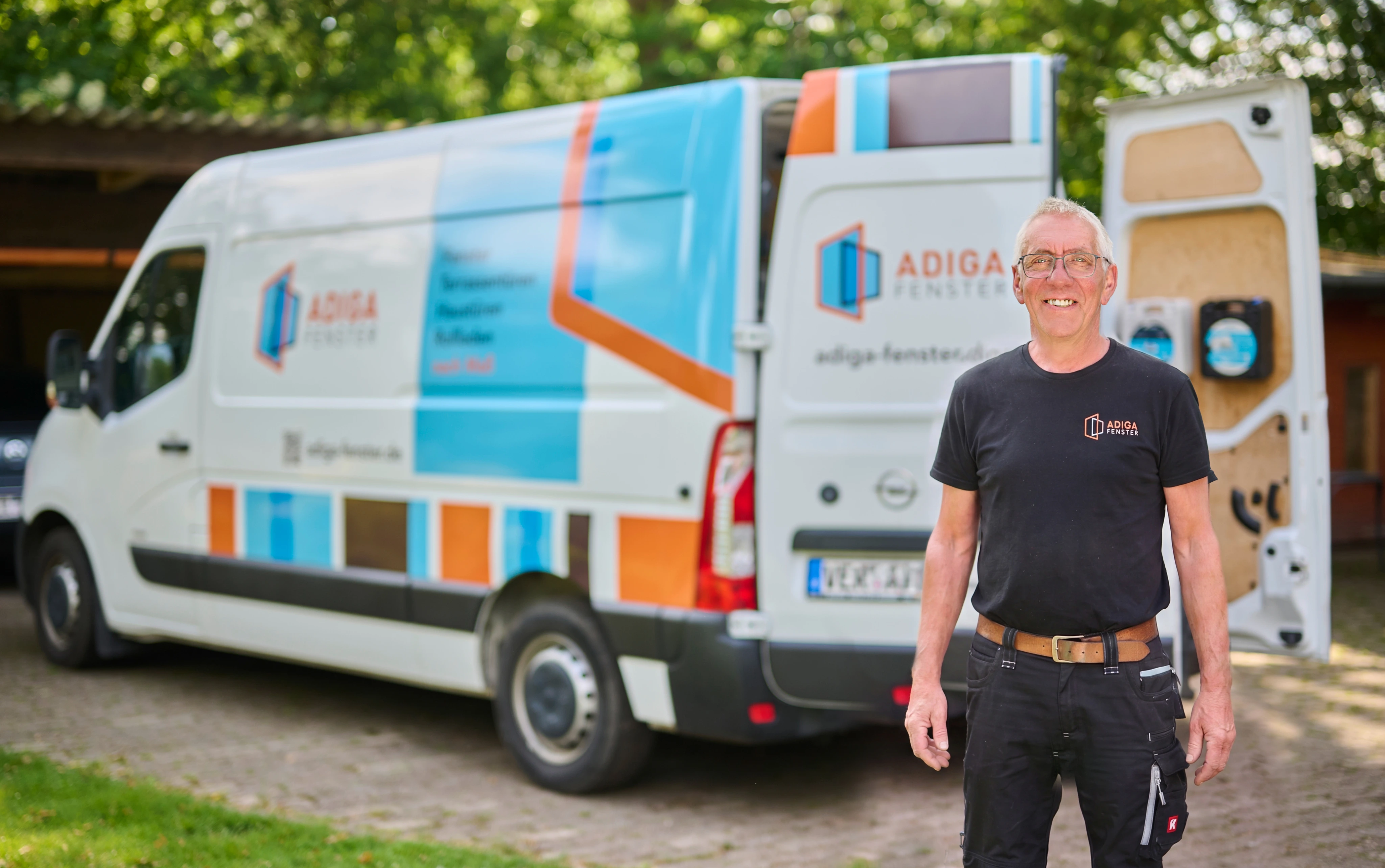 Ein lächelnder Mann steht vor einem Adiga-Fenster-Service-Van mit geöffneter Tür.