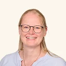 Frau mit blonden Haaren, Brille und lila Oberteil lächelnd vor hellem Hintergrund.