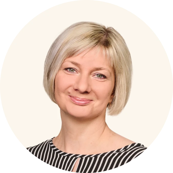 Porträt einer lächelnden Frau mit kurzem blondem Haar und gestreiftem Oberteil vor hellem Hintergrund.