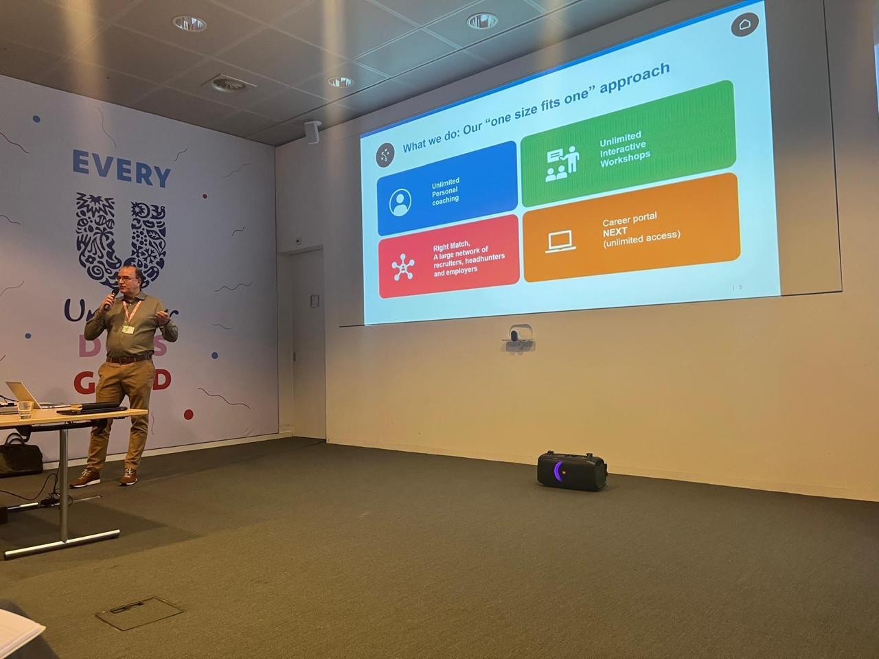 Adriaan stands next to a projection screen in a conference room, presenting a slide about a “one size fits one” approach with four colored sections.