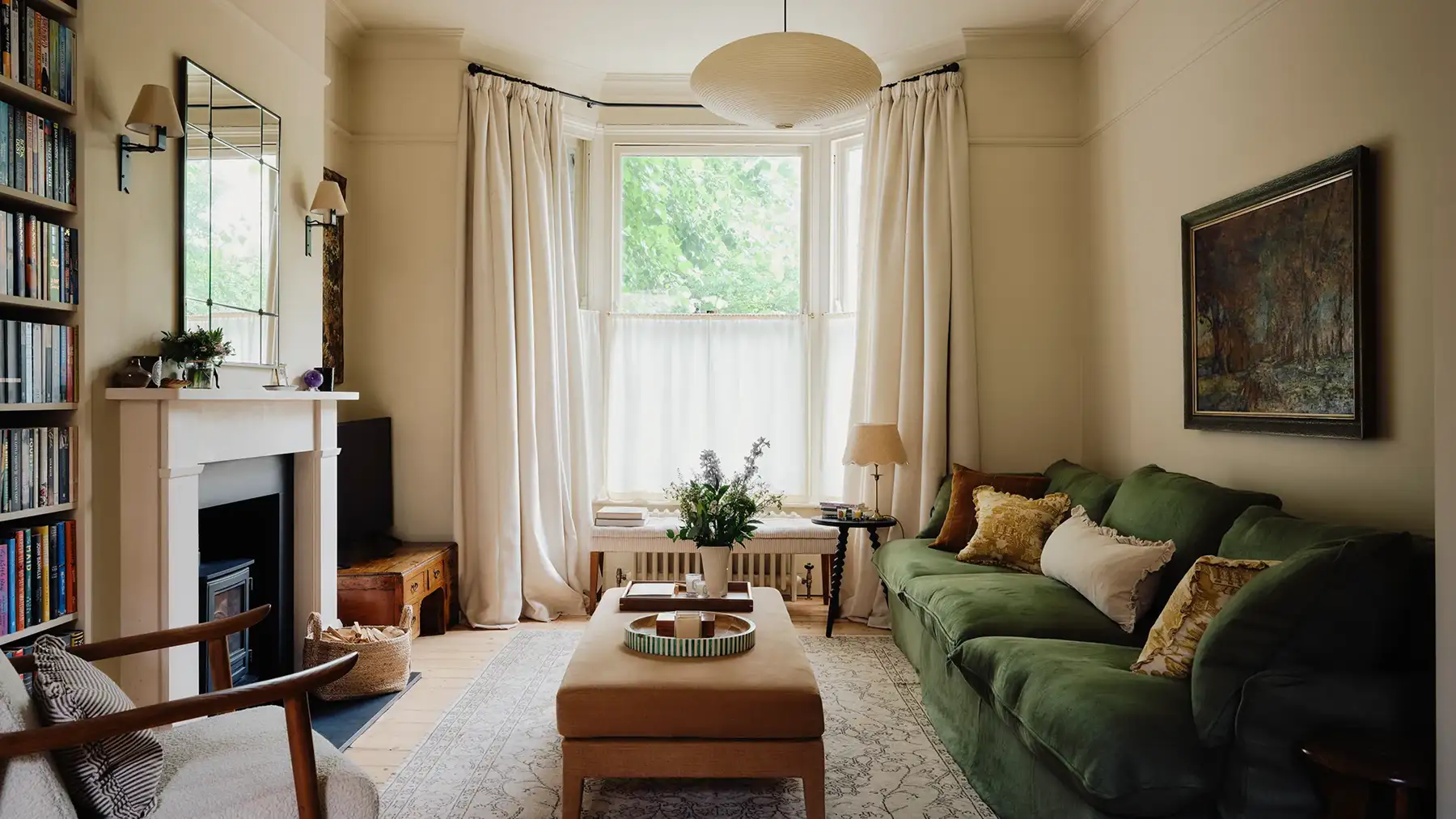 Cozy living room with green sofa, beige ottoman, fireplace, bookshelf, and large window with curtains.