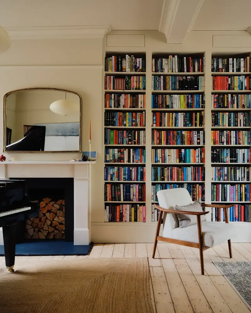 Living room with built-in bookshelves, a white armchair, a piano, fireplace, and a large mirror above the mantle.