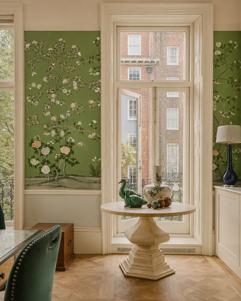 Elegant room with floral wallpaper, round table with ceramic decor, and large window overlooking buildings.