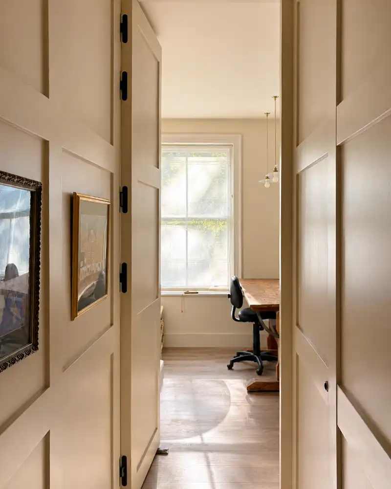 View through beige double doors into a room with a wooden desk, chair, and window with soft light.