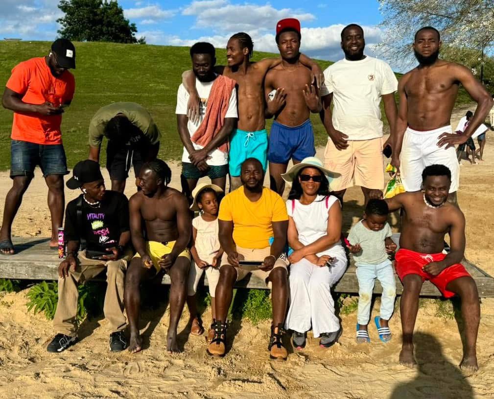 Group of men, one woman, and two children posing outdoors on sandy ground with a grassy hill and blue sky.