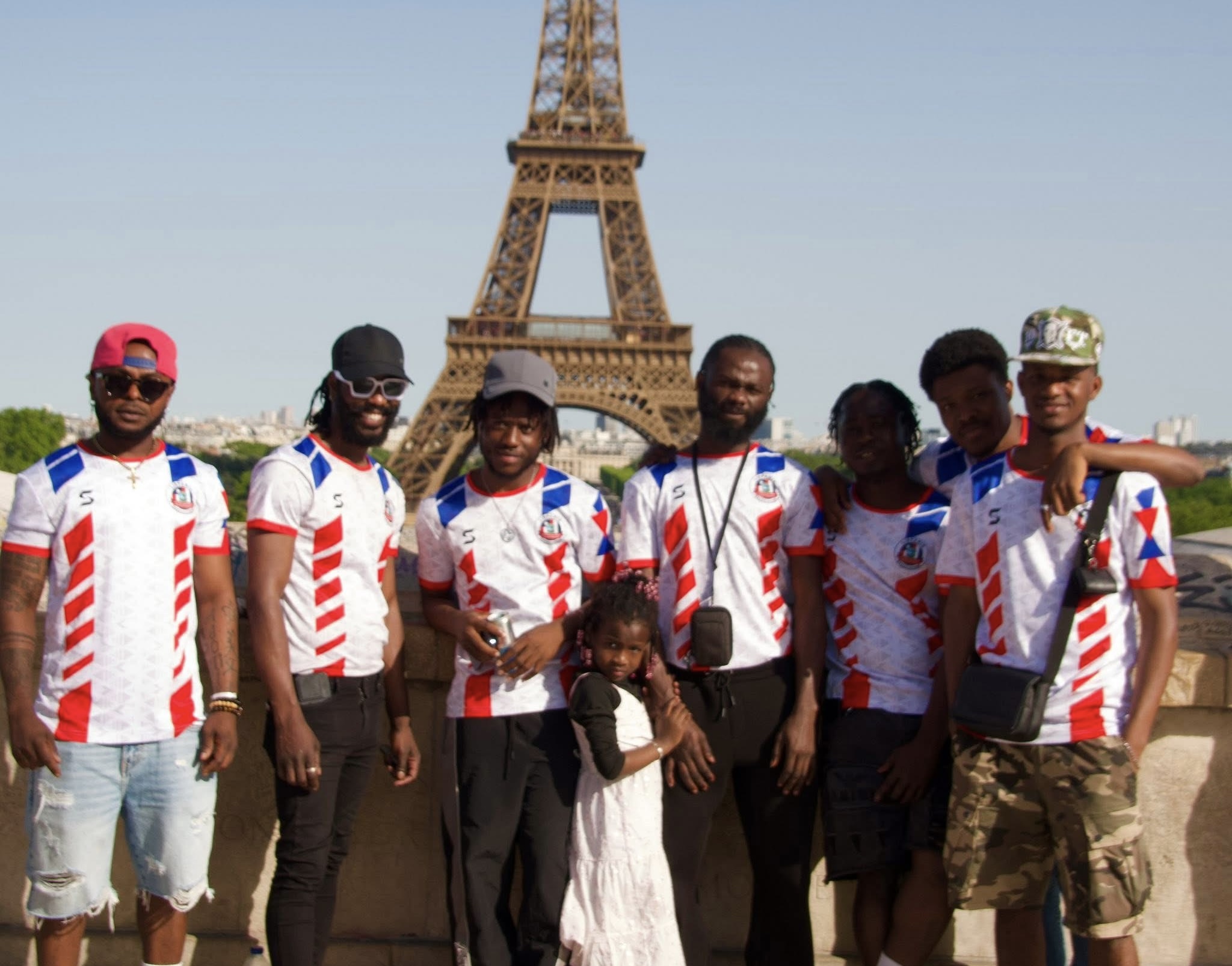 Group of men and a young girl posing in matching sports jerseys in front of the Eiffel Tower in daytime.