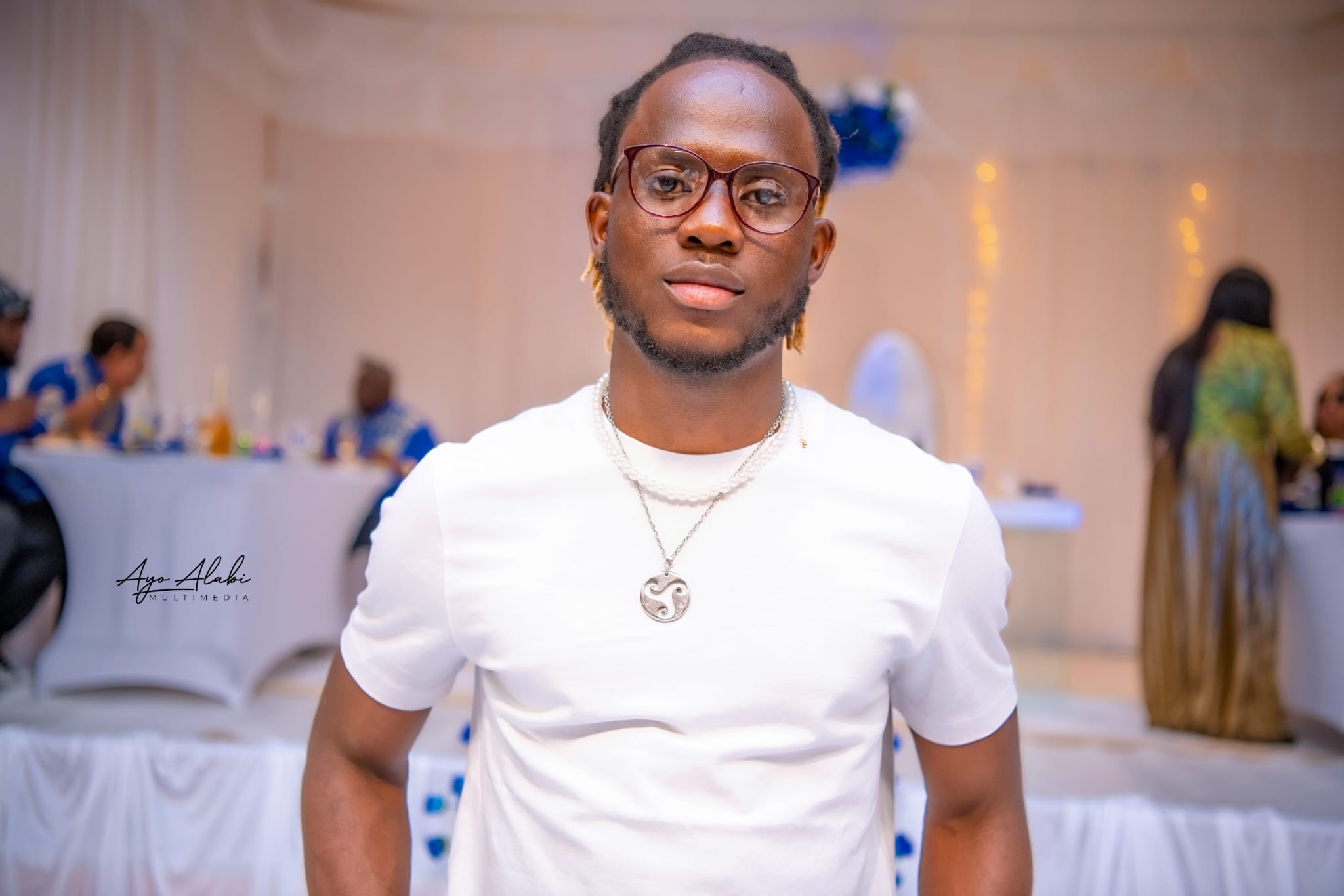 Man with glasses, beard, and dreadlocks wearing white shirt and pendant at an indoor event.