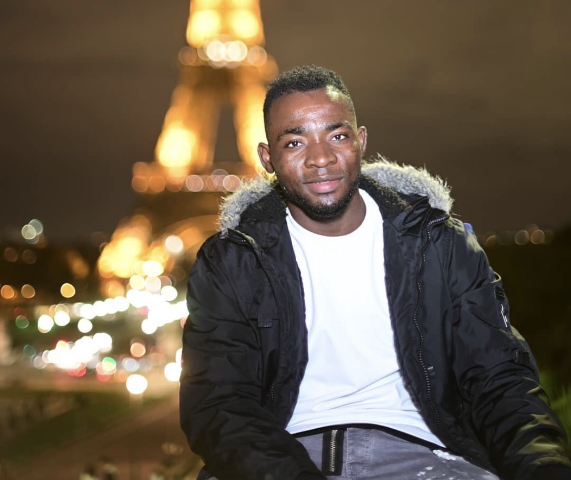 Smiling young man in black jacket and white shirt with blurred illuminated Eiffel Tower at night behind him.