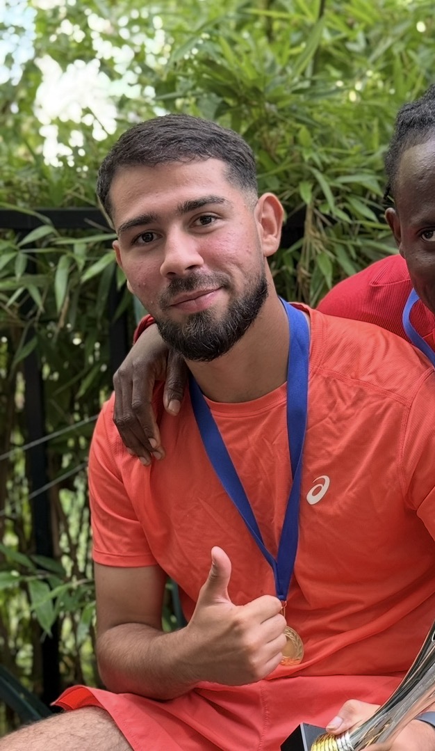 Man in orange shirt with medal around neck giving thumbs up, partially hugged by another person.