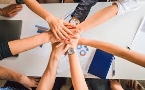 Multiple hands stacked together over a desk with office supplies and laptop nearby.