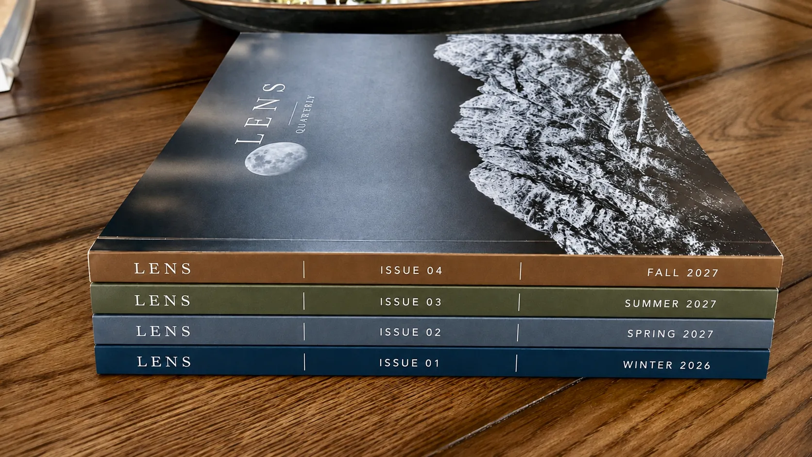 Four issues of LENS stacked on a wooden table, spines reading Winter 2026, Spring 2027, Summer 2027, and Fall 2027.