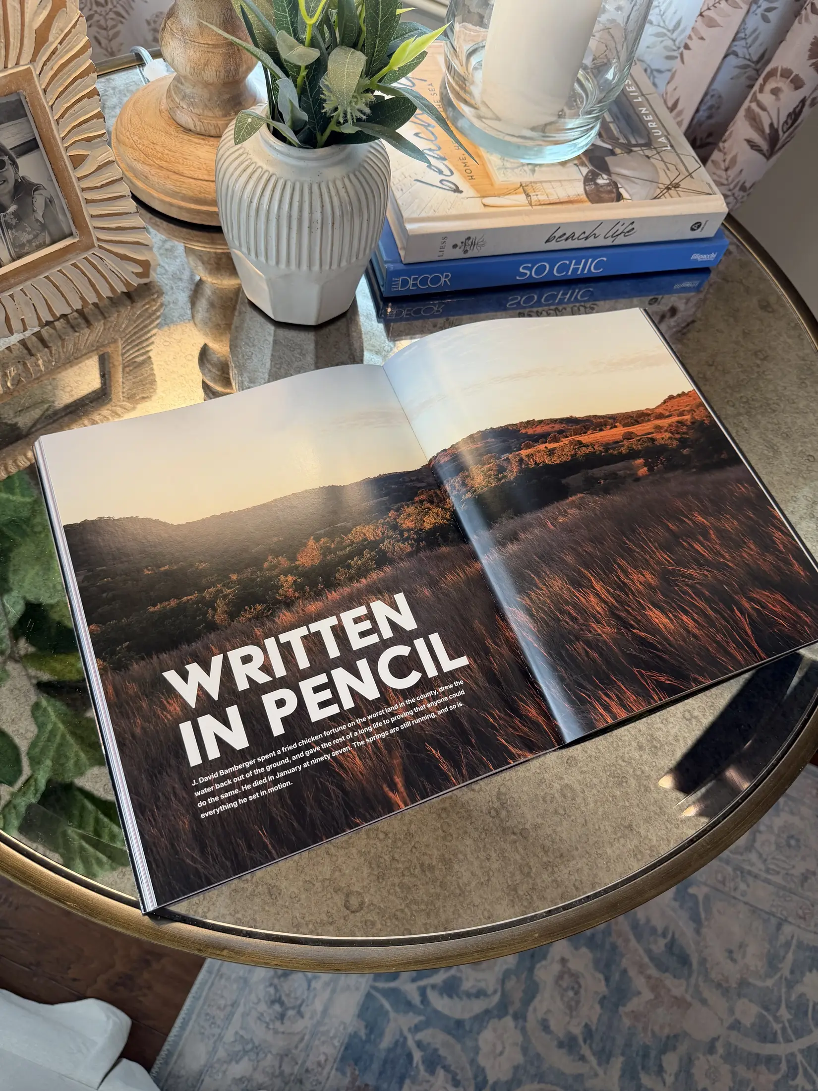 An issue of LENS open on a wooden table beside a stack of design books and a pair of reading glasses, the spread showing a vintage Bronco in long grass.