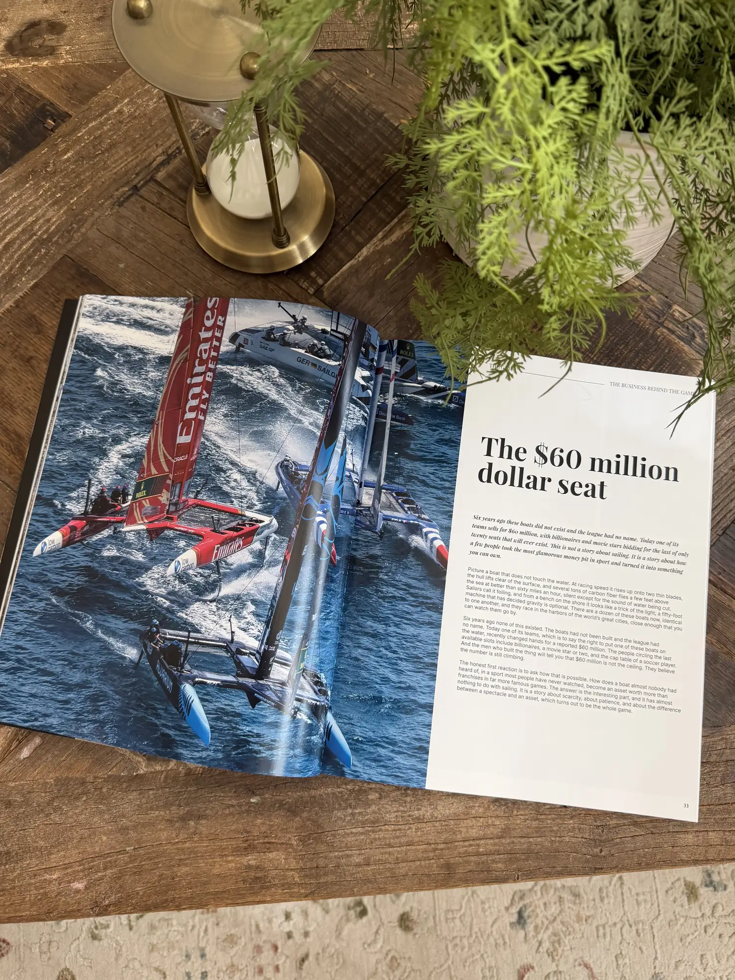 An issue of LENS lying open on a coffee table at a feature spread: racing catamarans across the left page, the headline The sixty million dollar seat on the right.